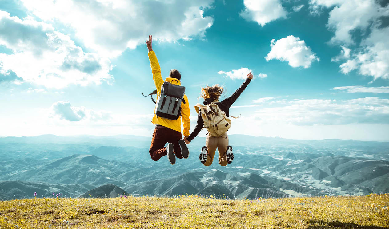 Hikers with backpacks jumping with arms up on top of a mountain - Couple of young happy travelers climbing the peak - Family, travel and adventure concept