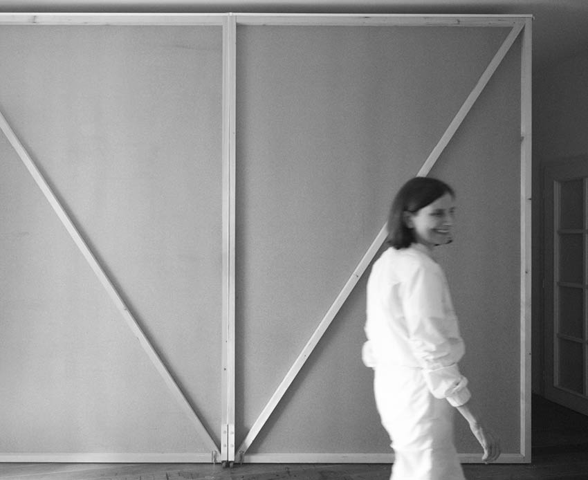 Oseba v svetlem obla ilu se premika mimo velikega lesenega okvirja z diagonalnima opornicama v minimalisti no opremljeni sobi. Description generated by AI